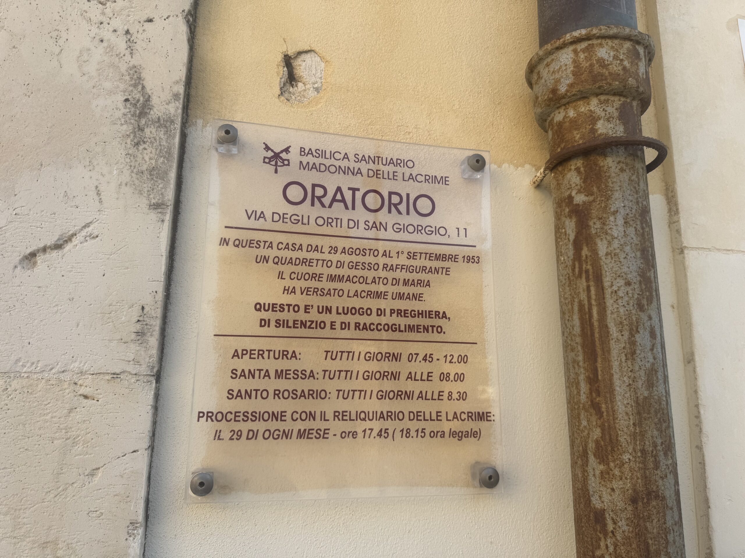Targa trasparente in plexiglass intitolata Basilica Santuario Madonna delle Lacrime – Oratorio, via degli Orti di San Giorgio 11, con il testo del prodigio e gli orari