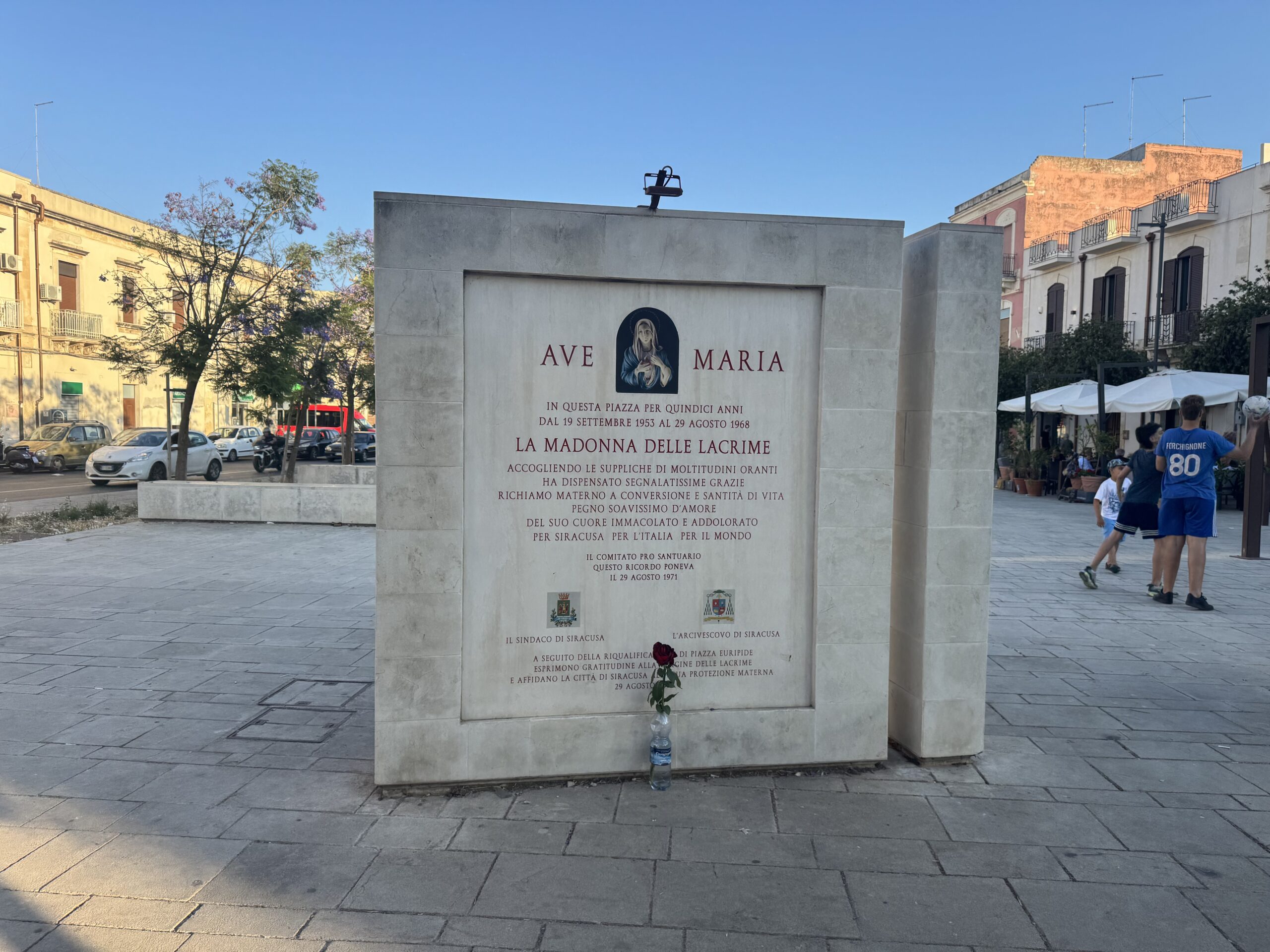 La stele vista di fronte con una rosa rossa deposta ai piedi e alcune persone nella piazza