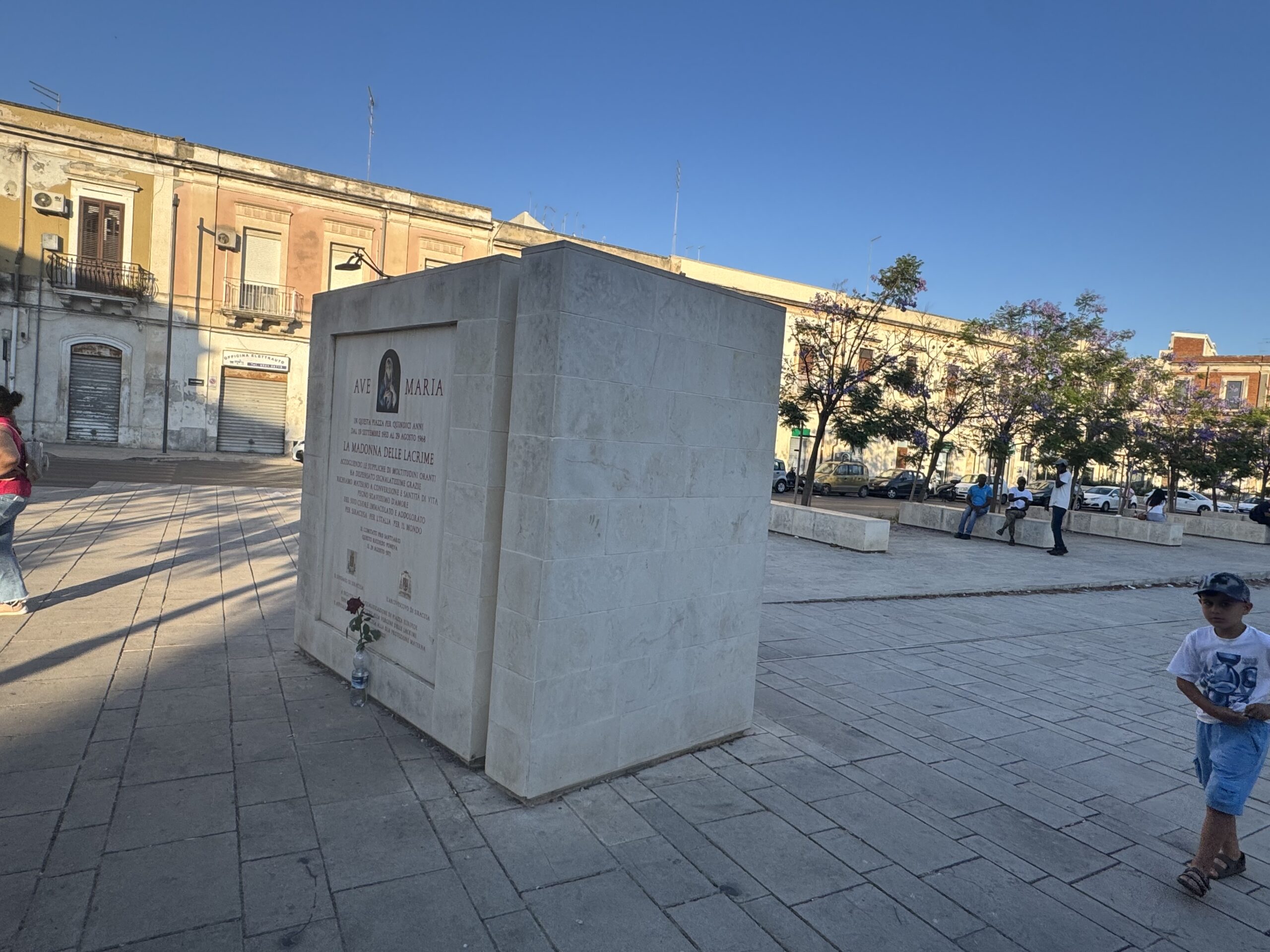 Monumento in lastre di pietra chiara a forma di libro aperto al centro di Piazza Euripide a Siracusa, con croce sulla sommità, alberi di jacaranda in fiore e palazzi della Borgata sullo sfondo