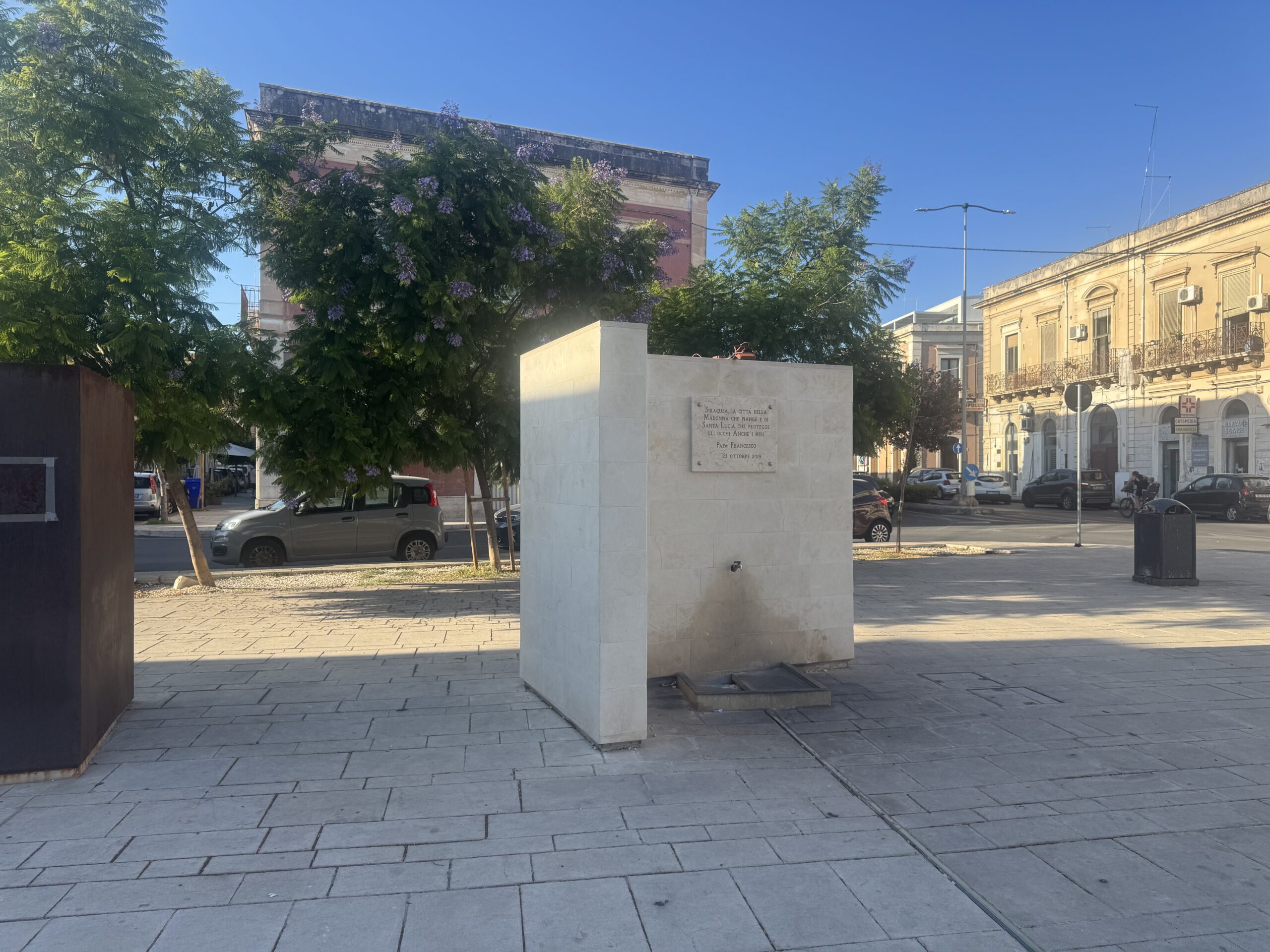 Il retro del monumento in pietra chiara nella piazza, con la lapide di papa Francesco, alberi di jacaranda e palazzi