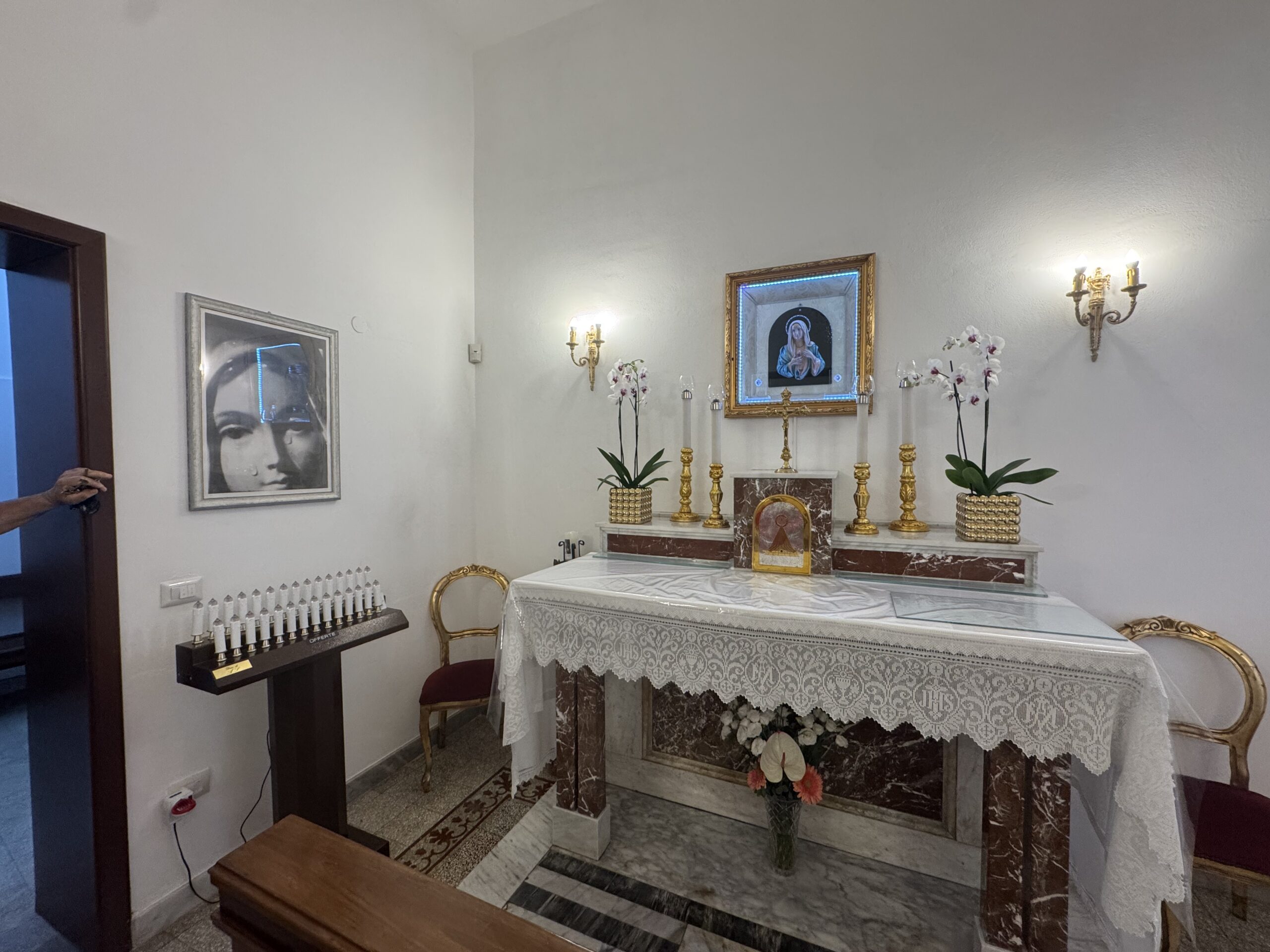 Parete dell'oratorio con una grande riproduzione fotografica in bianco e nero del volto della Madonna del quadretto e, accanto, un espositore di ceri votivi