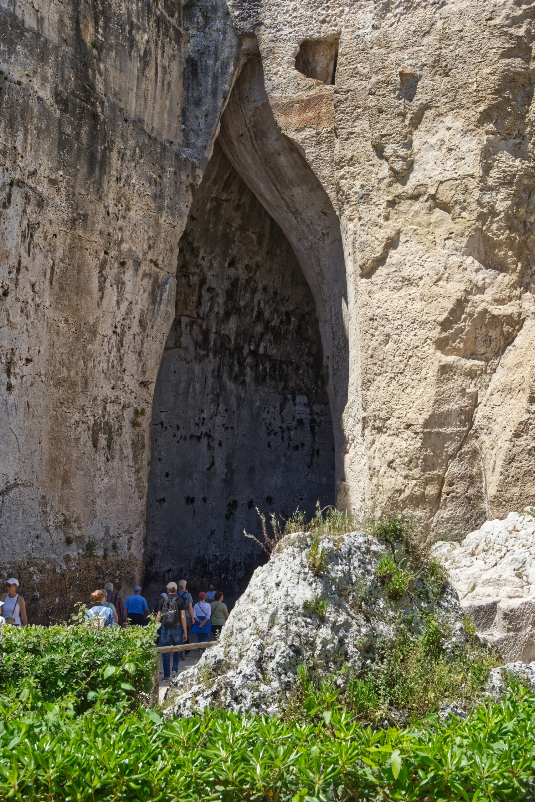 La fessura verticale altissima dell'ingresso dell'Orecchio di Dionisio, con le pareti di roccia che si curvano e si stringono verso l'alto e un gruppo di visitatori alla base che ne mostra la scala