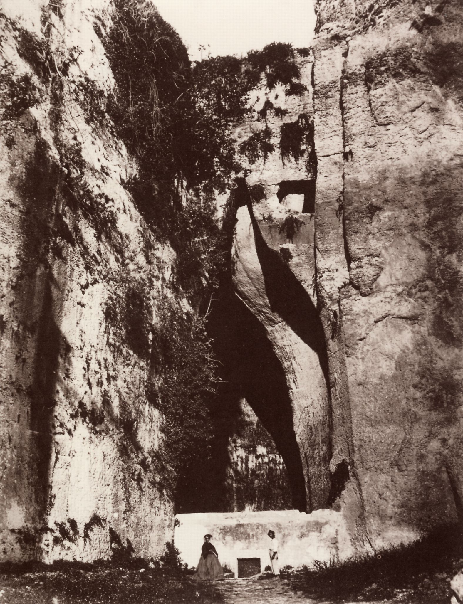 Fotografia ottocentesca in bianco e nero: la fessura della grotta con l'ingresso chiuso da un muro bianco con porta, davanti una coppia in abiti d'epoca con la donna in crinolina