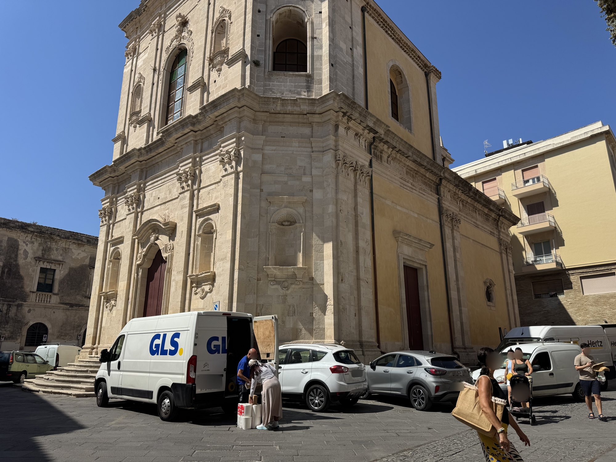 Chiesa barocca isolata al centro di una piazza, con la facciata in pietra chiara a sinistra e il lungo fianco intonacato di ocra a destra, davanti auto e furgoni in sosta