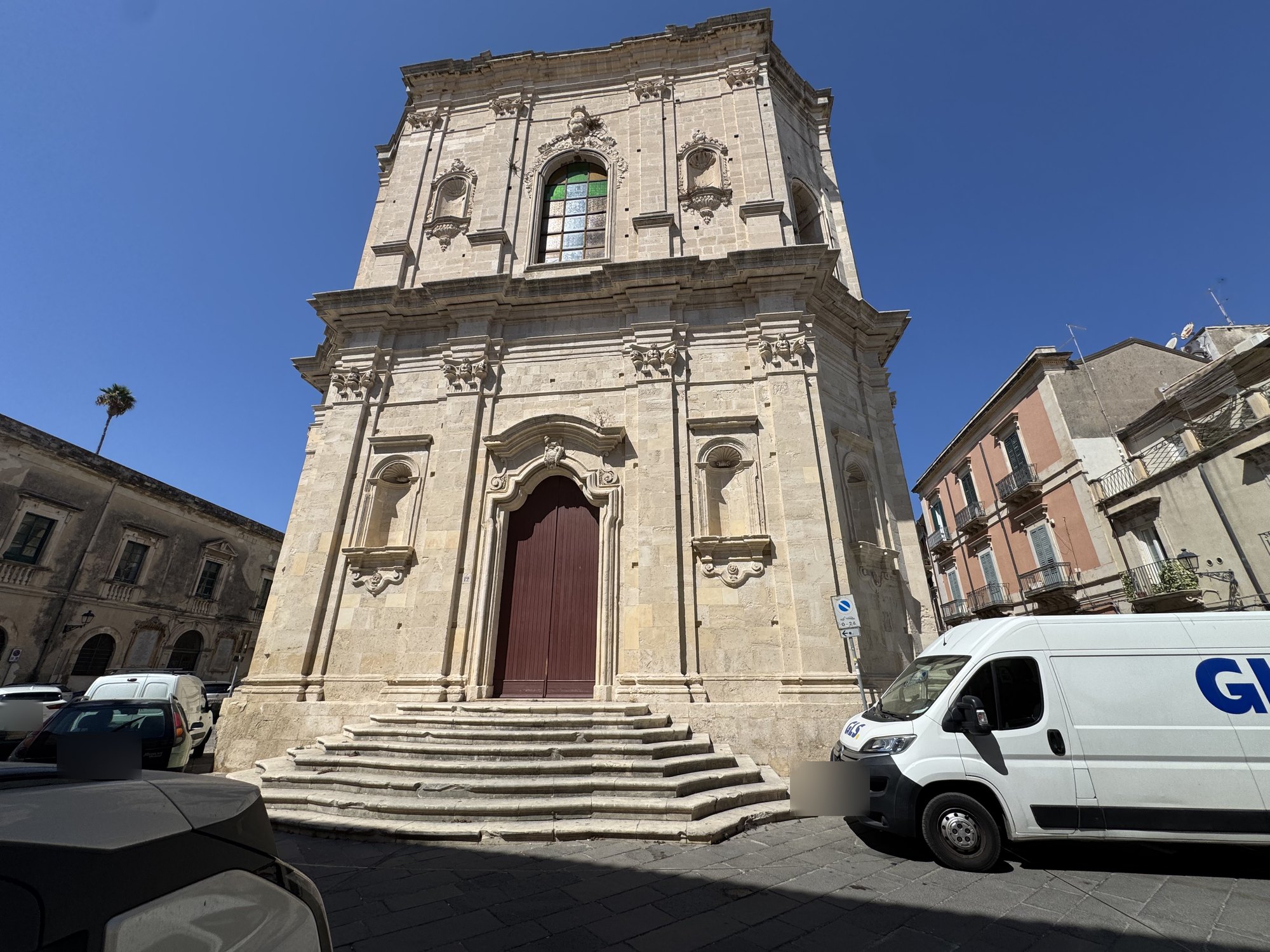 Facciata barocca in pietra chiara su due ordini con portale color bordeaux e scalinata curvilinea di gradini in pietra che sale dalla piazza