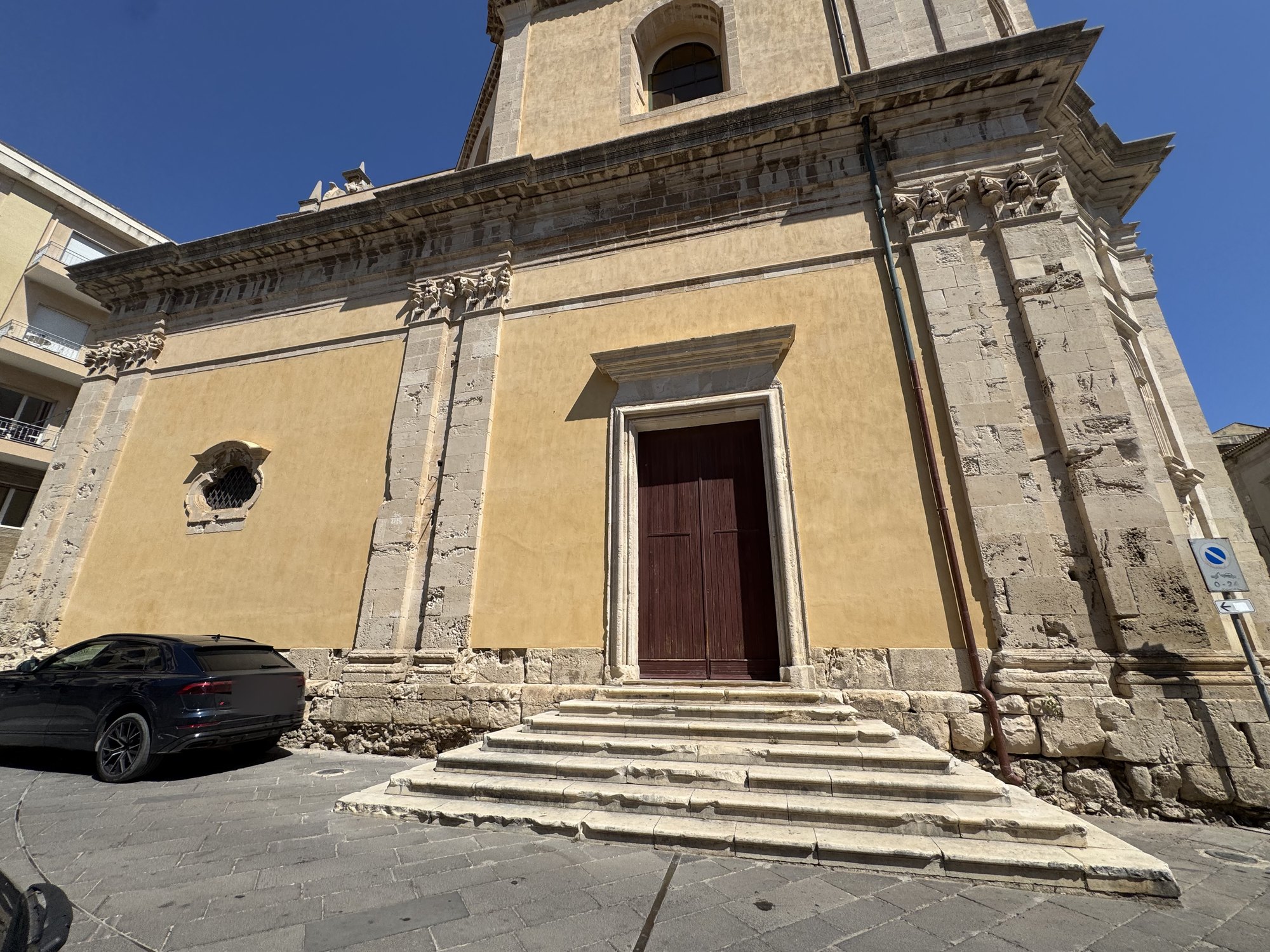 Portale laterale con cornice in pietra preceduto da cinque gradini e, sulla parete, una piccola finestra ovale con grata