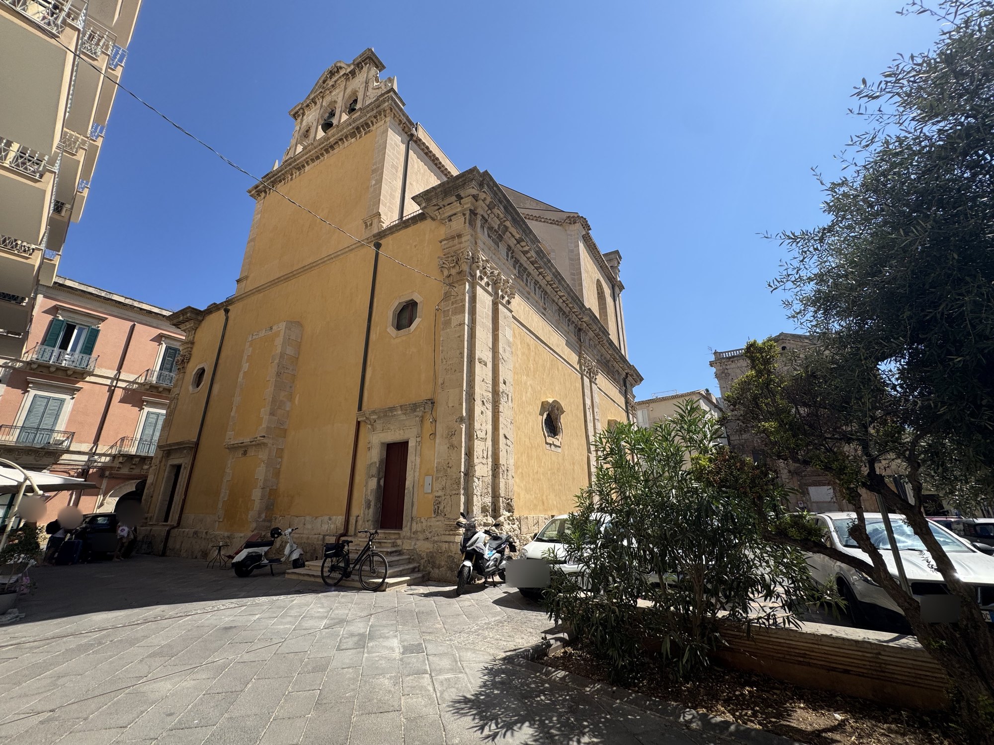 Retro della chiesa con campanile a vela in pietra a due fornici che sormonta la parete intonacata di ocra, davanti una piccola aiuola con ulivo e oleandri