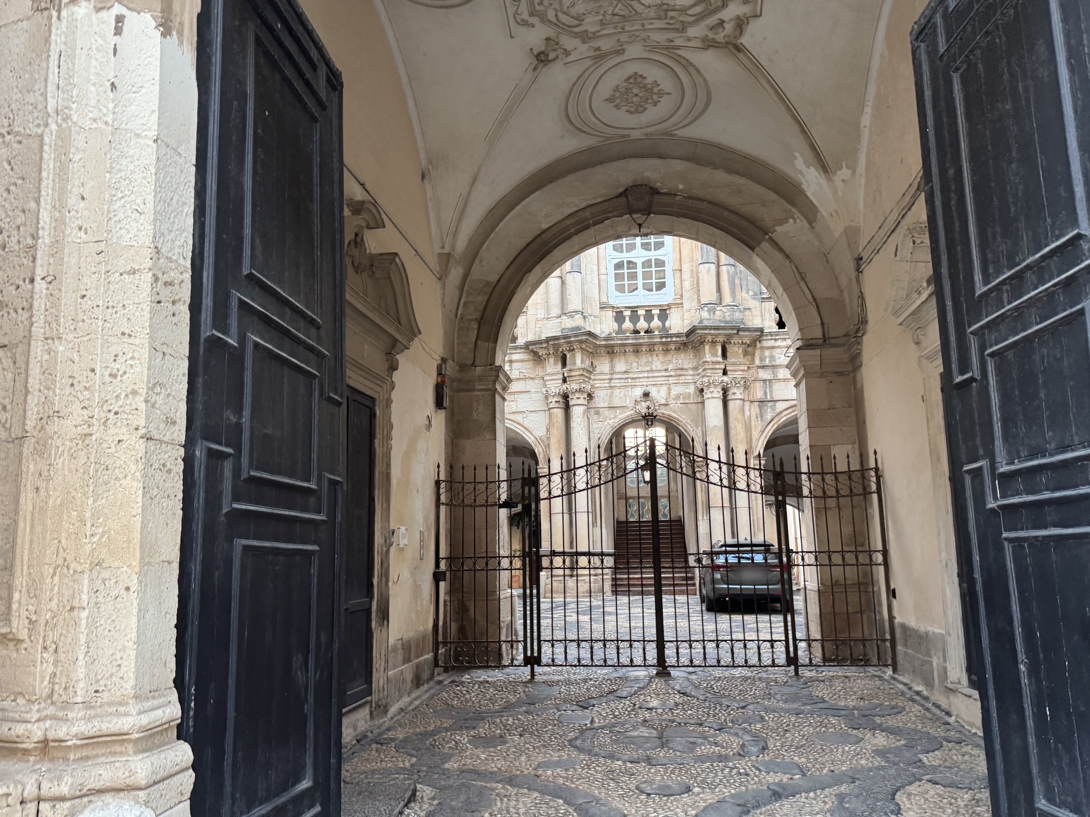 Androne coperto da volta con unghie e stucchi, pavimentato a ciottoli disposti a disegno, chiuso in fondo da una cancellata in ferro oltre la quale si vedono il cortile e una scalinata