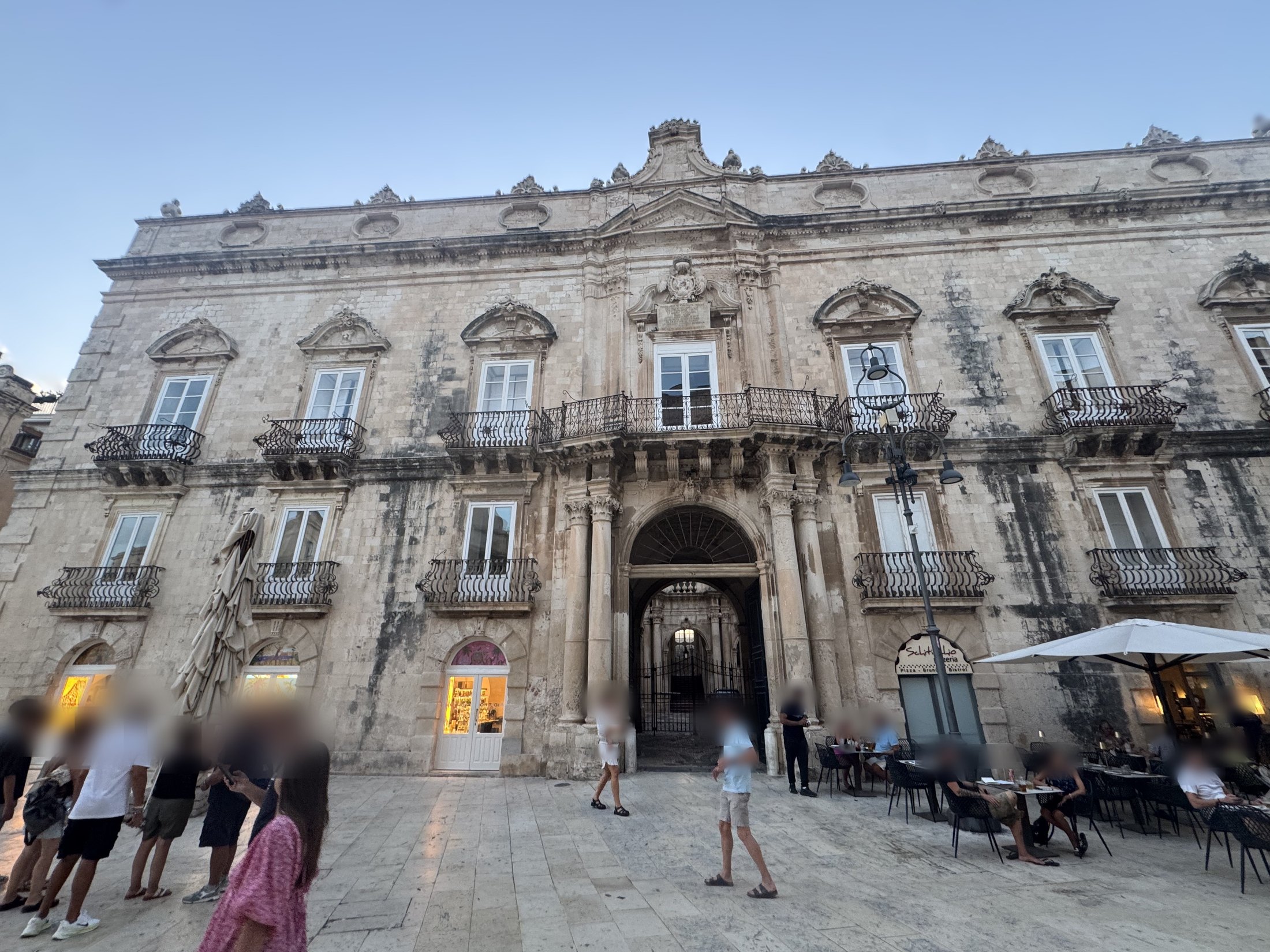 Veduta d'insieme della facciata del palazzo dalla piazza al tramonto, con passanti e tavolini di un locale davanti all'edificio