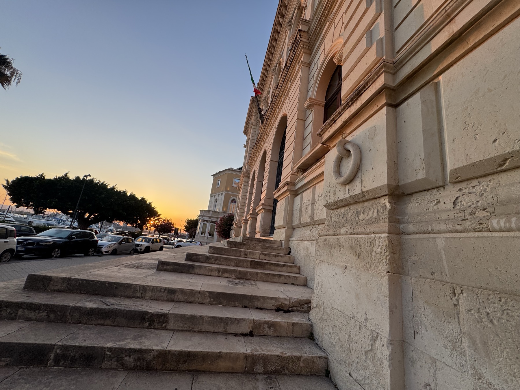 Dettaglio del basamento in pietra con un grande anello scolpito in rilievo, sullo sfondo il porto al tramonto