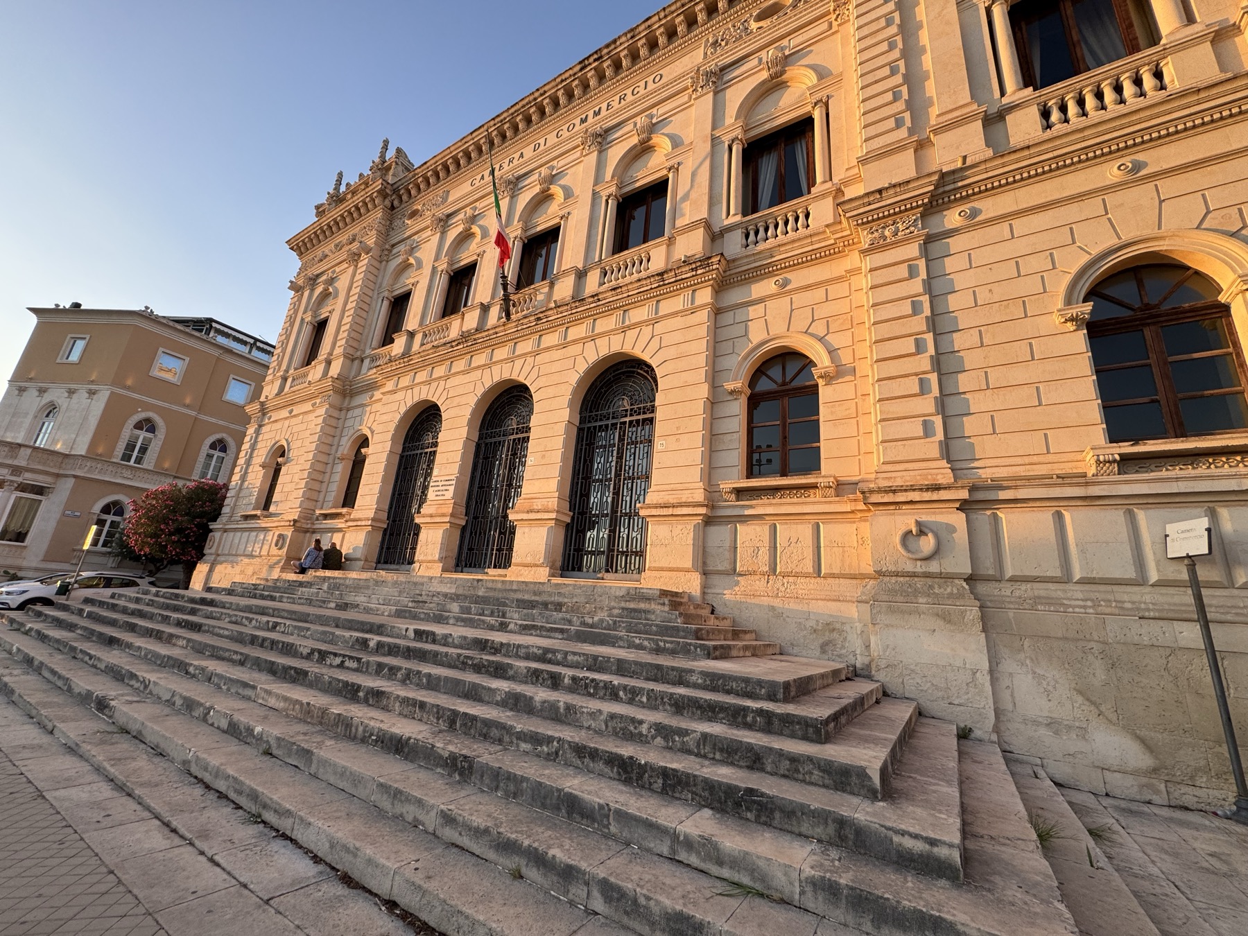 Scorcio del palazzo dal basso con la scalinata monumentale, i tre portali ad arco chiusi da cancellate e il fregio con la scritta CAMERA DI COMMERCIO