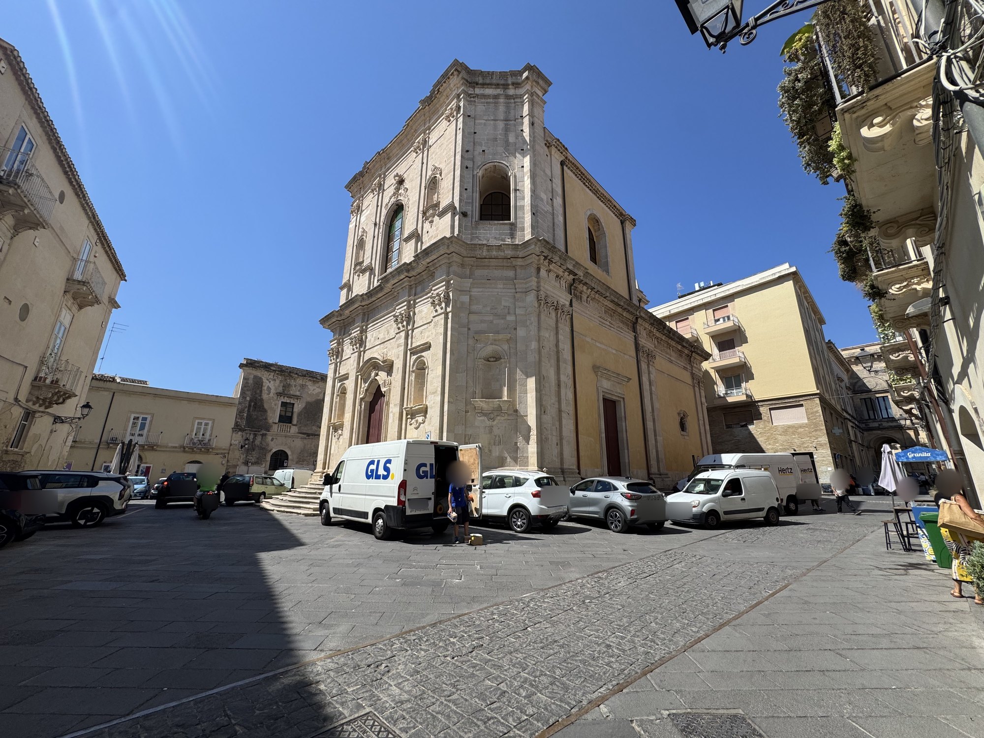Ampia piazza pavimentata in basole scure con al centro la chiesa barocca isolata, palazzi storici sui lati e auto in sosta