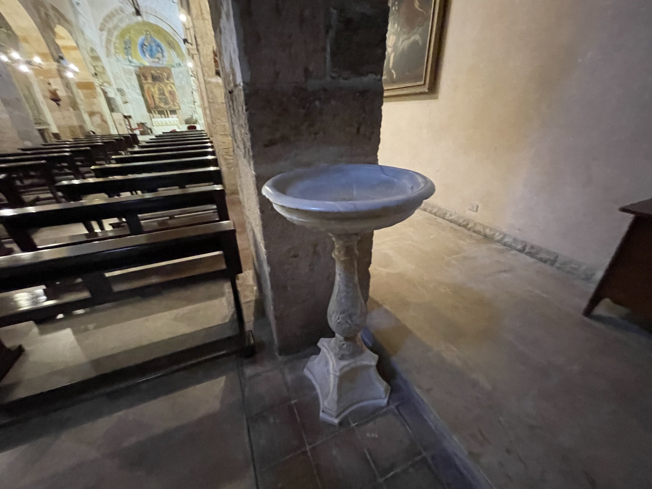 L'acquasantiera di marmo accanto al pilastro, con i banchi di legno e la navata sullo sfondo