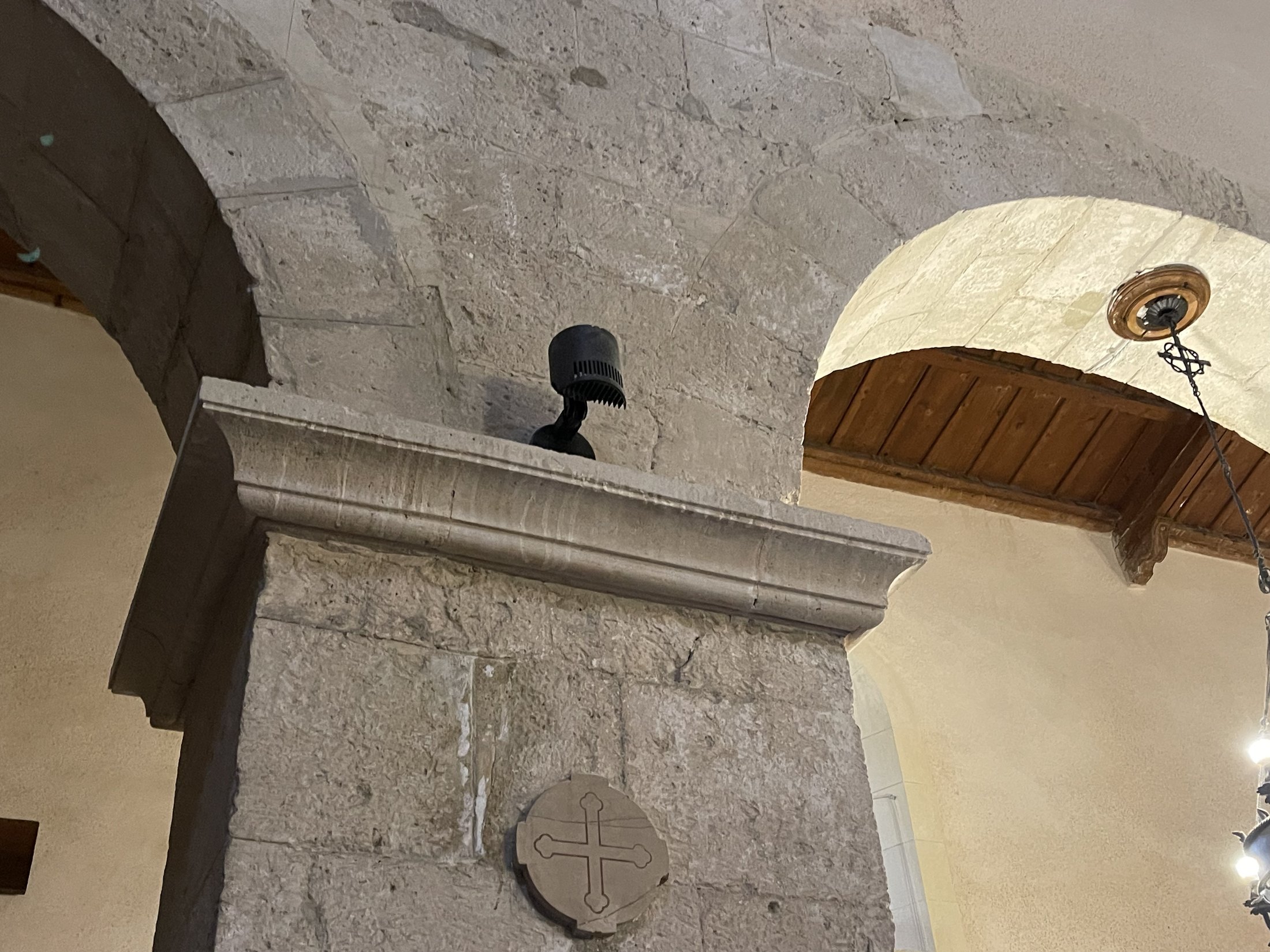 Croce greca scolpita entro un disco di pietra sotto la cornice d'imposta di un pilastro, con il soffitto ligneo sullo sfondo