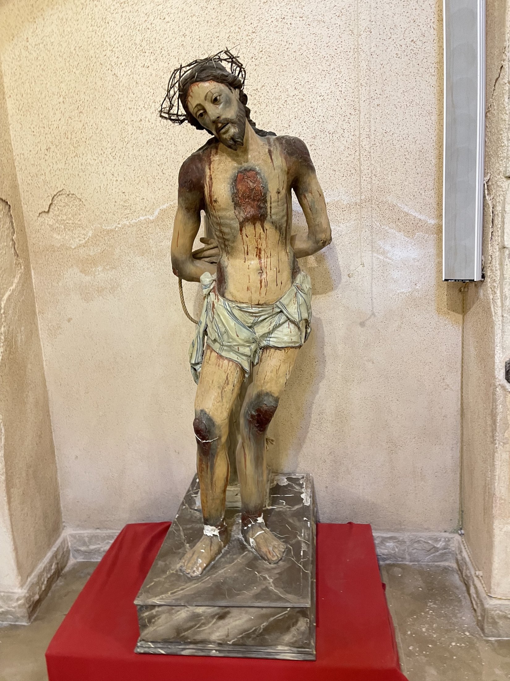 Statua policroma a grandezza naturale dell'Ecce Homo, con corona di spine, perizoma e piaghe, su una base coperta di panno rosso