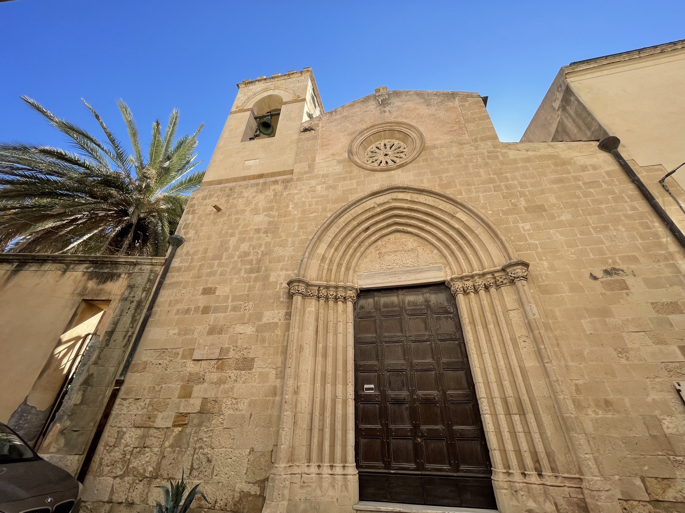 La facciata in conci di calcarenite della chiesa di San Martino vista di scorcio, con il portale ogivale, il rosone e il campanile a torre sulla sinistra, fra le fronde di una palma