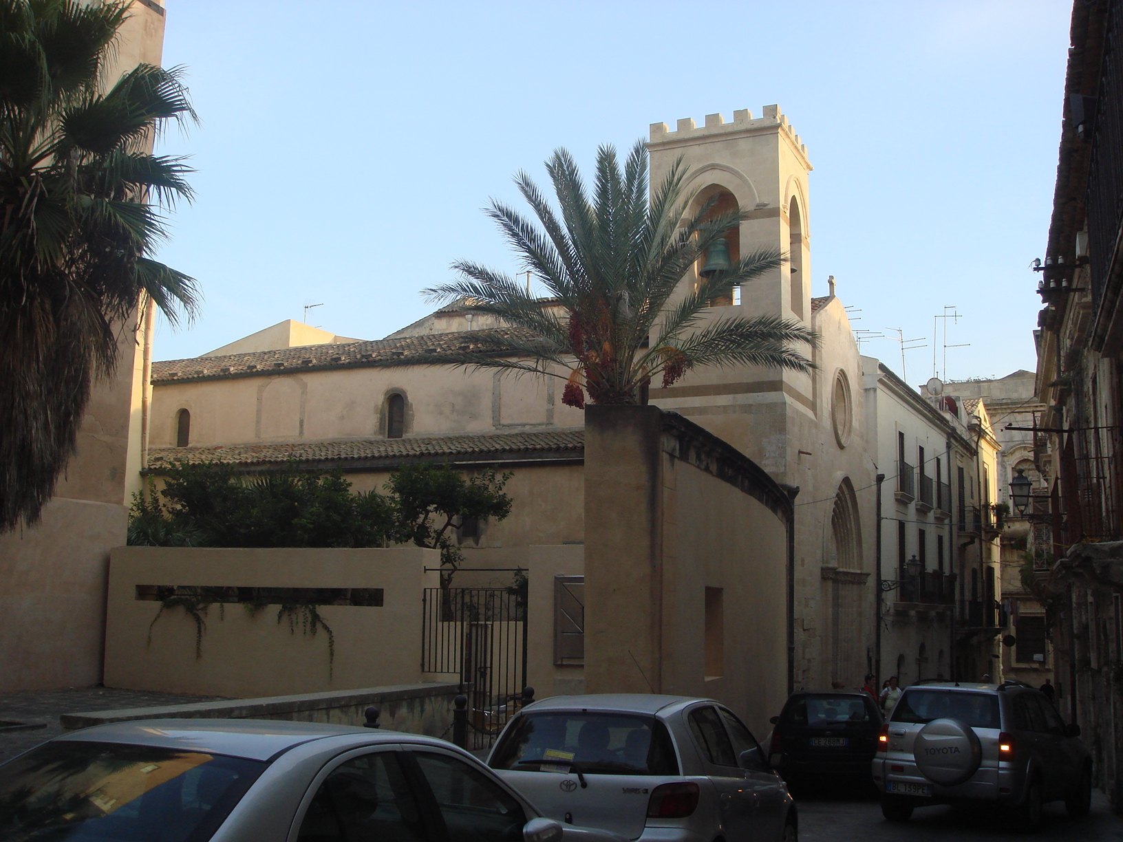 Il fianco nord della chiesa oltre il muro del cortile: il tetto a coppi della navatella, il muro alto della navata con le monofore e le sagome dei finestroni tamponati, il campanile merlato