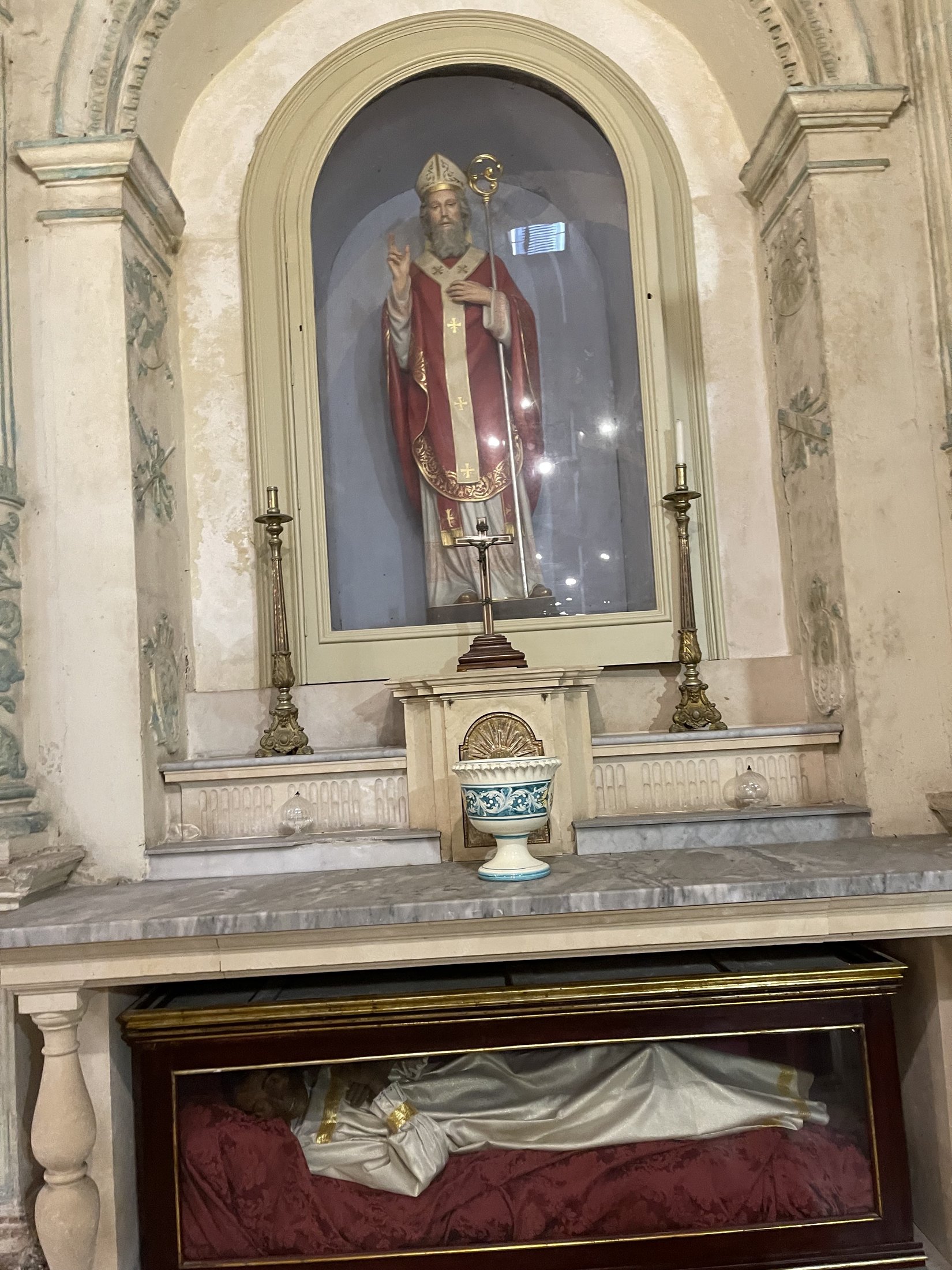 Statua policroma di un santo vescovo benedicente in abiti rossi, con mitra e pastorale, entro una nicchia ad arco con vetrina, fra due candelieri