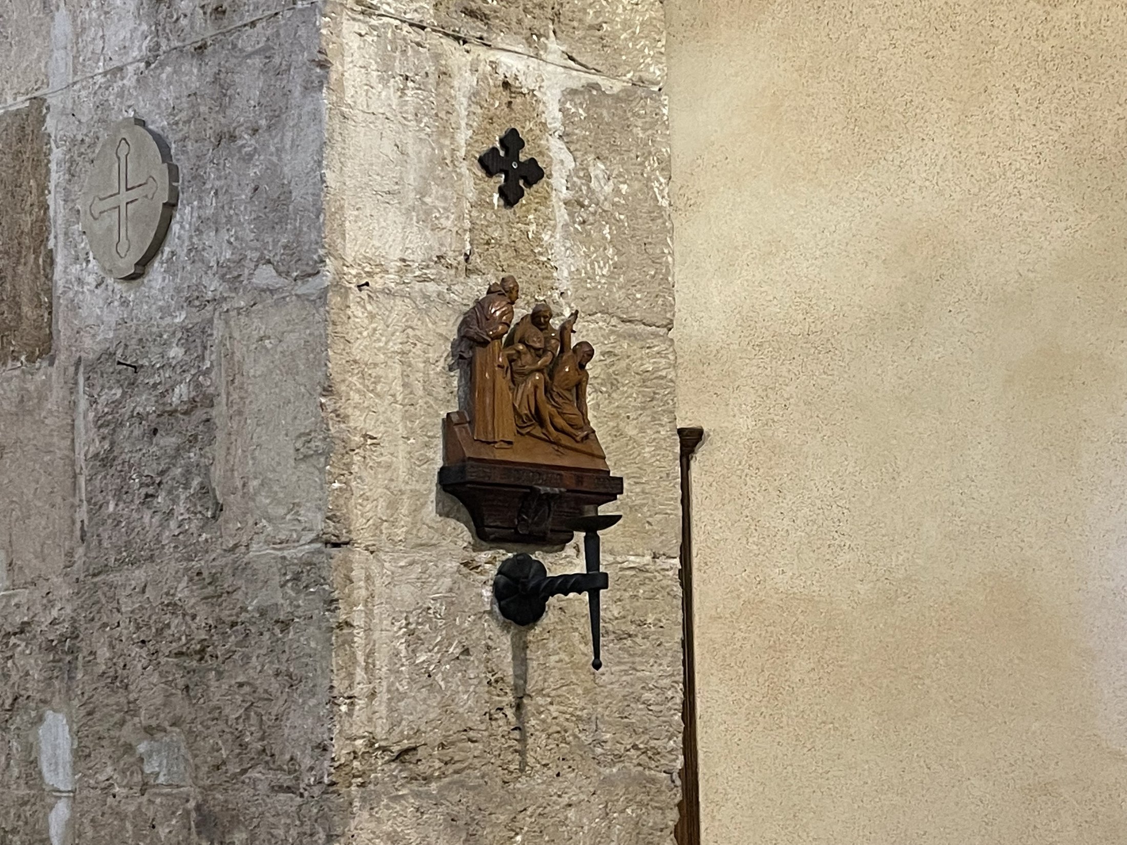 Pilastro in conci di pietra con una croce greca scolpita entro un cerchio e, accanto, una stazione lignea della Via Crucis su mensola