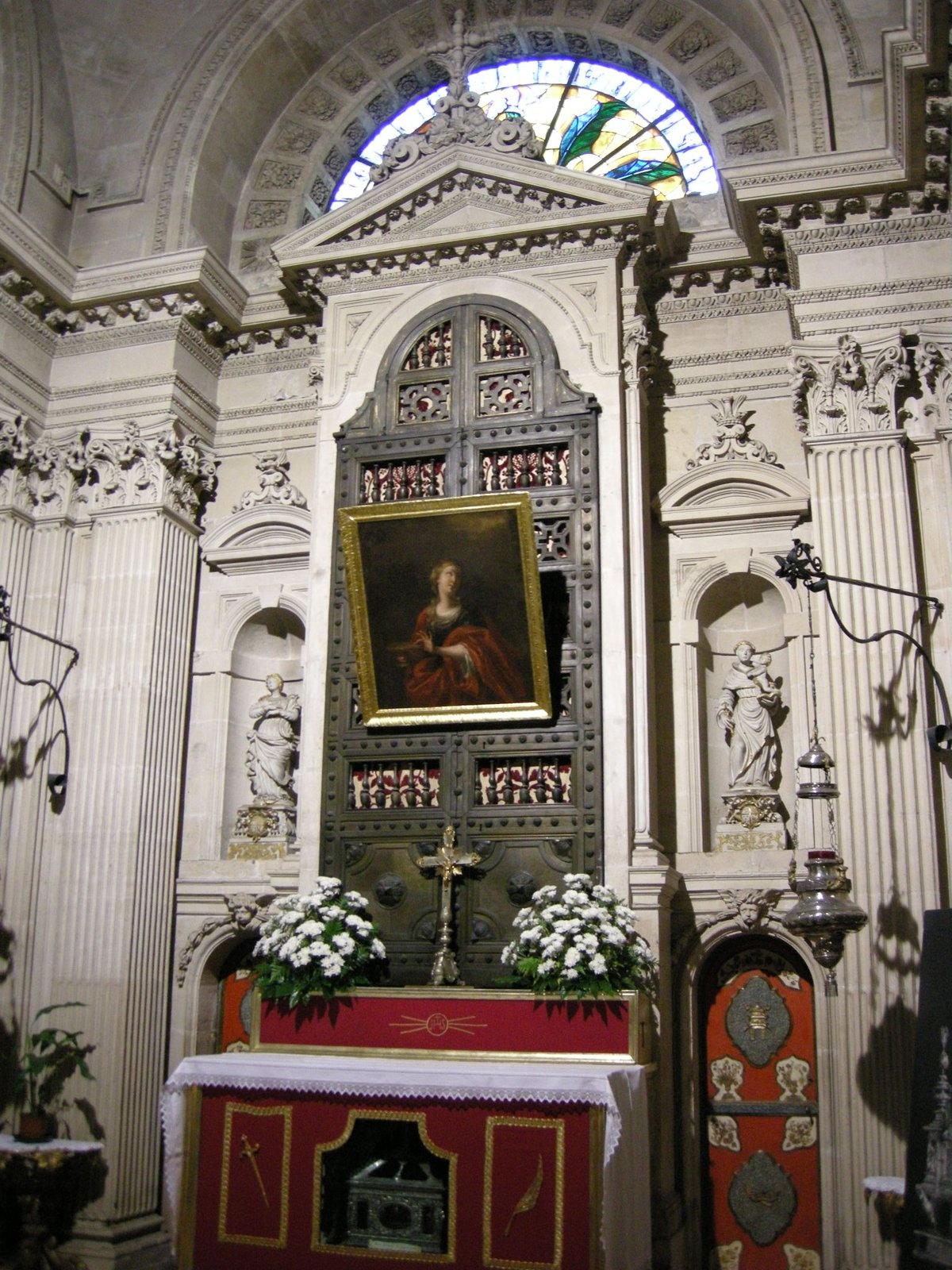 L'altare della Cappella di Santa Lucia nel Duomo di Siracusa: una nicchia chiusa da una grande porta metallica scura a due battenti, sormontata da un timpano marmoreo riccamente intagliato, con appeso un dipinto a olio della santa in veste rossa; sopra si apre una lunetta con vetrata colorata, ai lati due statuette entro nicchie, davanti l'altare rivestito di un paliotto rosso e oro con due vasi di fiori bianchi e un crocifisso.