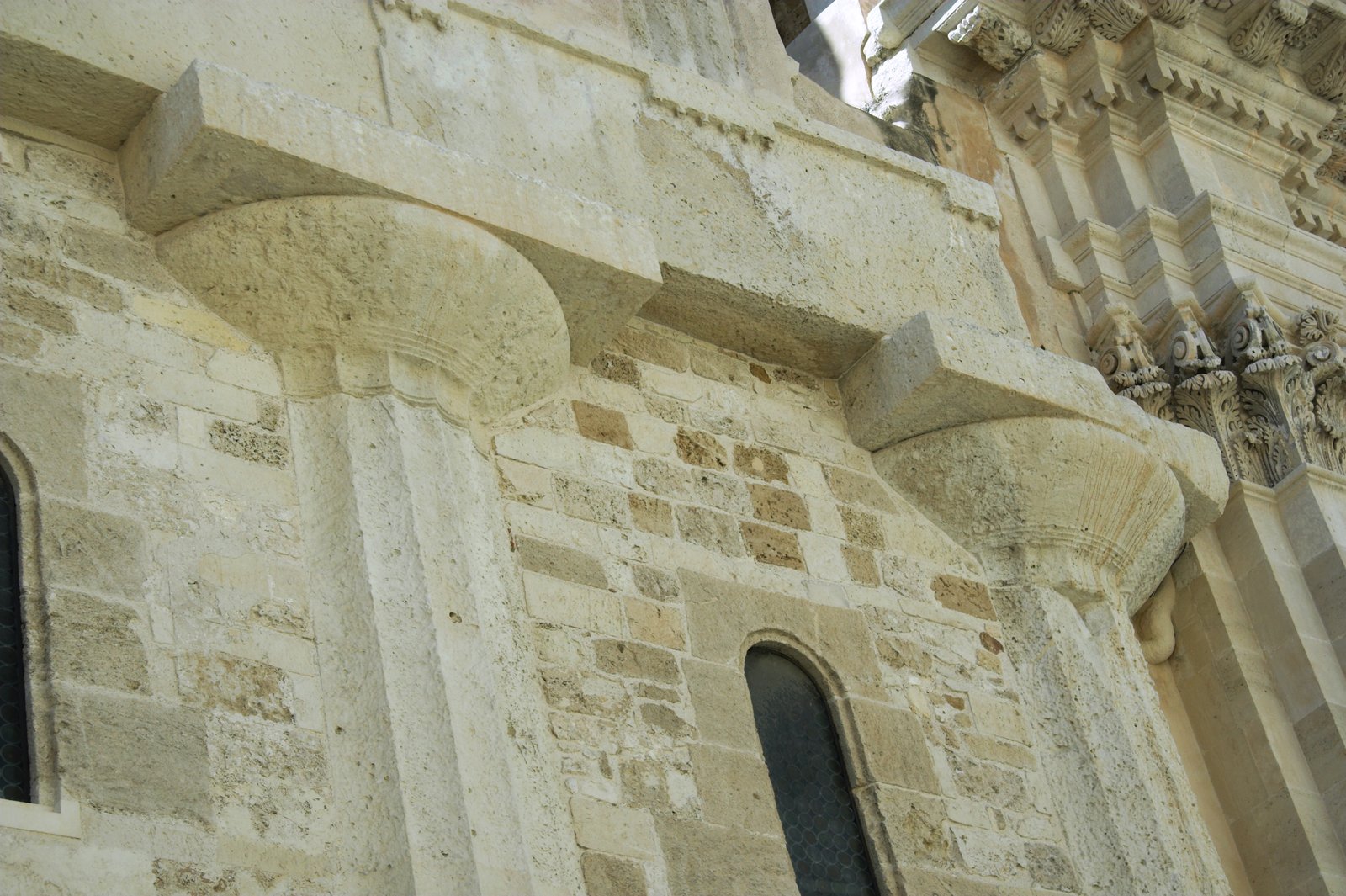 Dettaglio ravvicinato del muro esterno del Duomo di Siracusa: due capitelli dorici del tempio di Atena, con larghi echini a cuscino sotto abachi quadrati, emergono dalla muratura in conci di pietra che li ha inglobati; fra le colonne si aprono due strette finestre ad arco e in alto a destra si intravede la decorazione barocca della facciata.