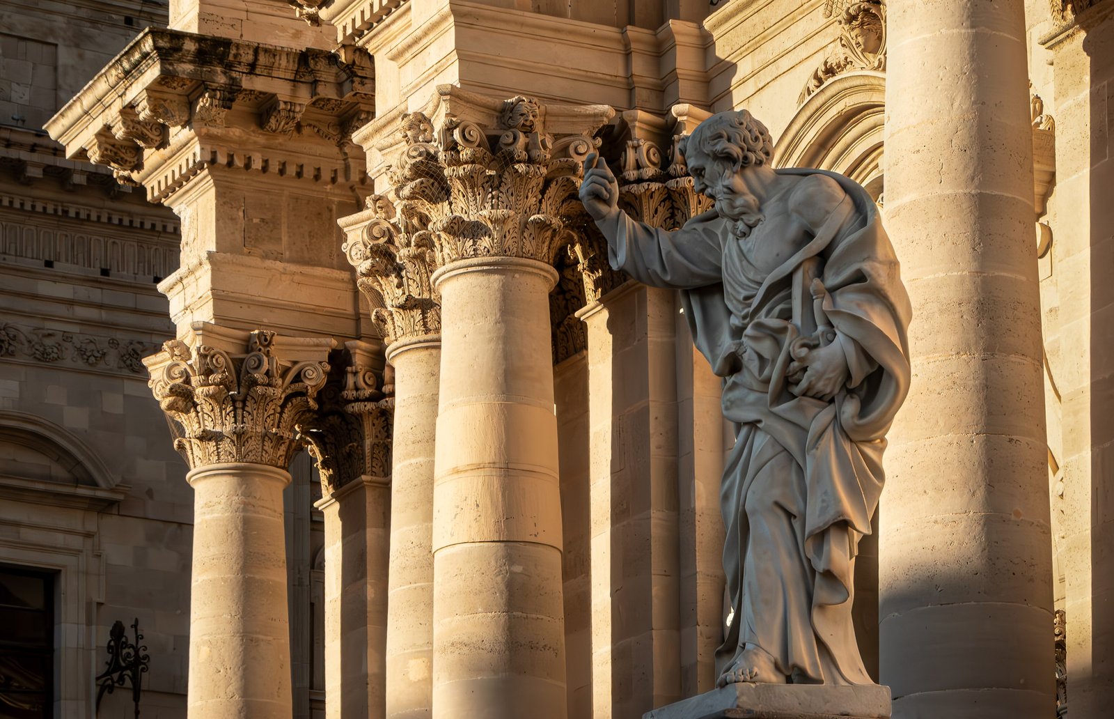 Scorcio ravvicinato della facciata barocca del Duomo di Siracusa ripresa dal basso nella luce dorata del tramonto: quattro colonne in pietra calcarea con capitelli corinzi a foglie d'acanto e volute, e in primo piano la statua marmorea di San Paolo con il braccio destro sollevato e l'ampio panneggio mosso.