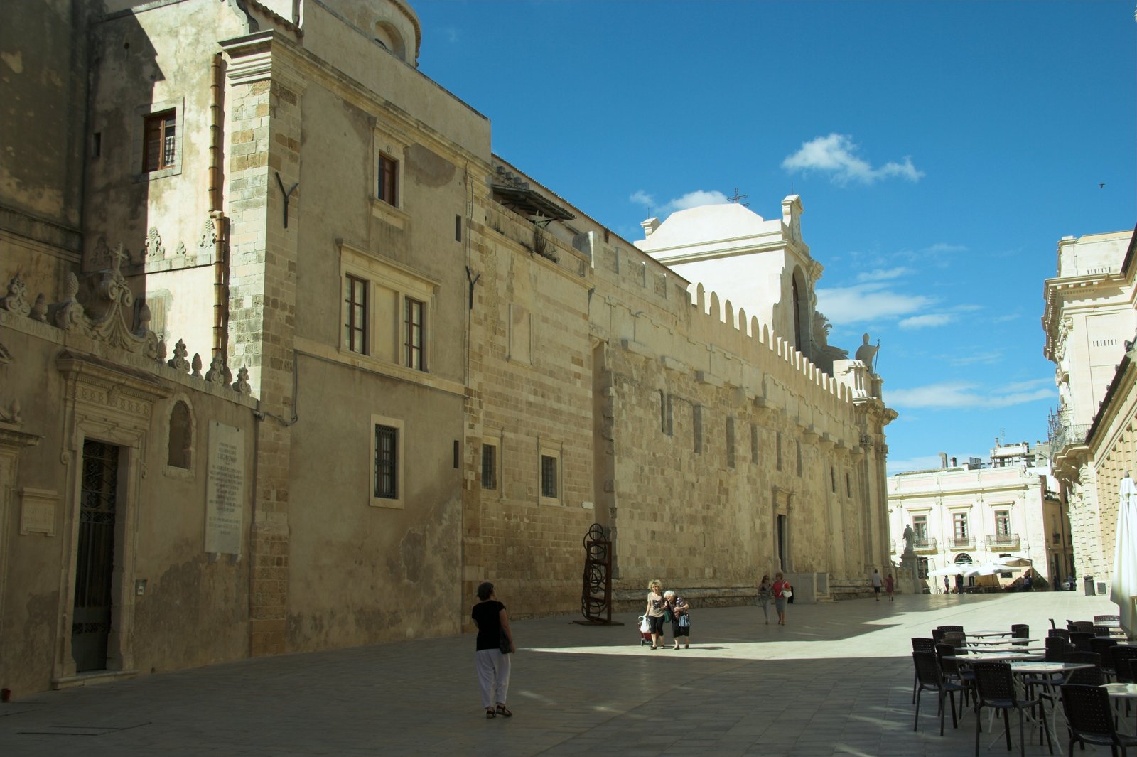 Il lungo fianco settentrionale del Duomo di Siracusa visto da piazza Duomo: muraglia in blocchi di calcare chiaro con piccole finestre rettangolari, coronamento a merli arrotondati e, sullo sfondo, il corpo bianco della cupola; in primo piano la piazza lastricata con alcuni passanti e i tavolini di un bar.