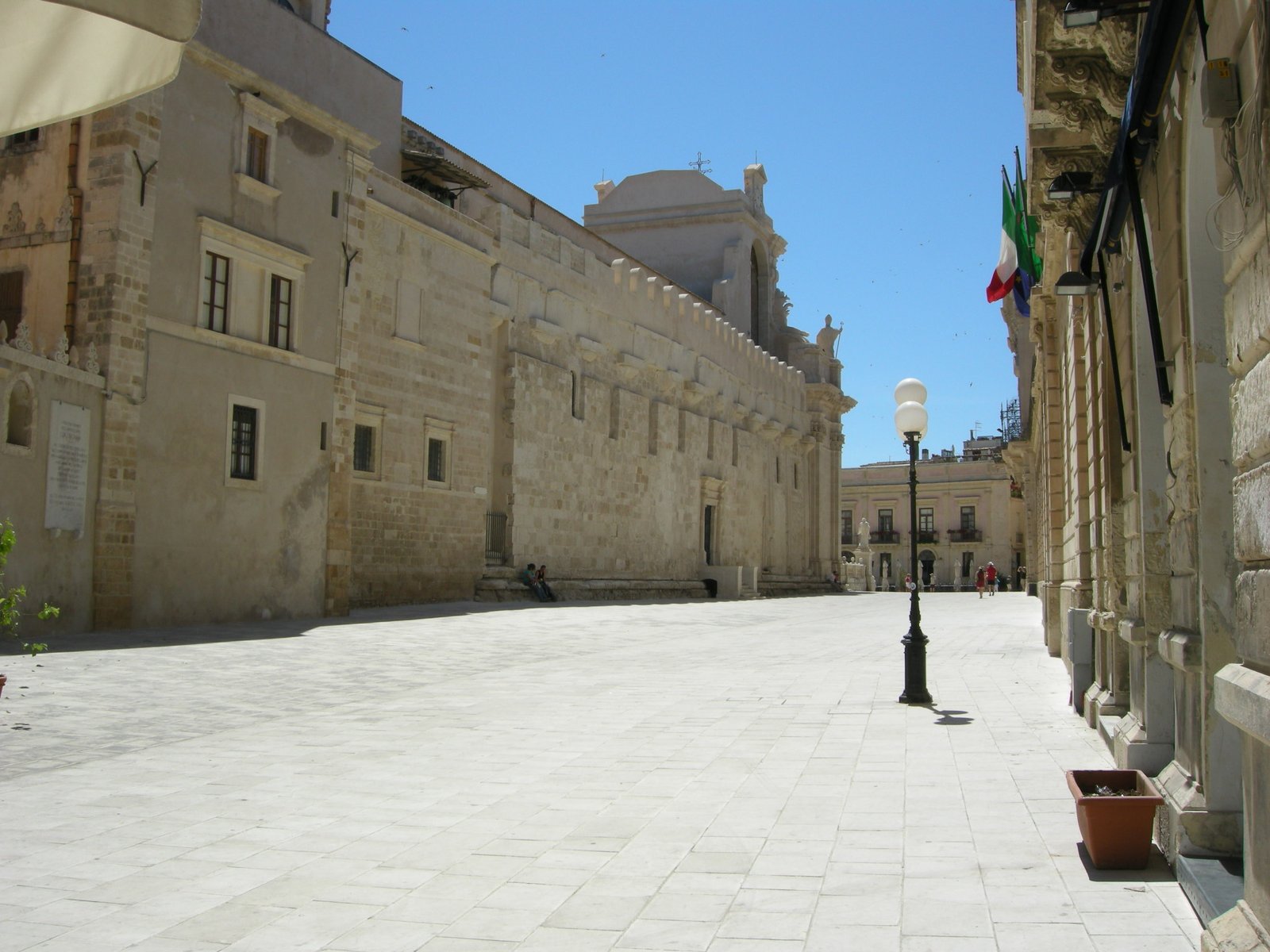 Veduta di via Minerva a Ortigia con il fianco del Duomo di Siracusa sulla sinistra: muro in conci di pietra chiara con coronamento a merli, la cupola chiara che emerge dietro il tetto e la strada lastricata in pietra bianca che scende verso piazza Duomo, con un lampione in ghisa e alcune bandiere sulla destra.