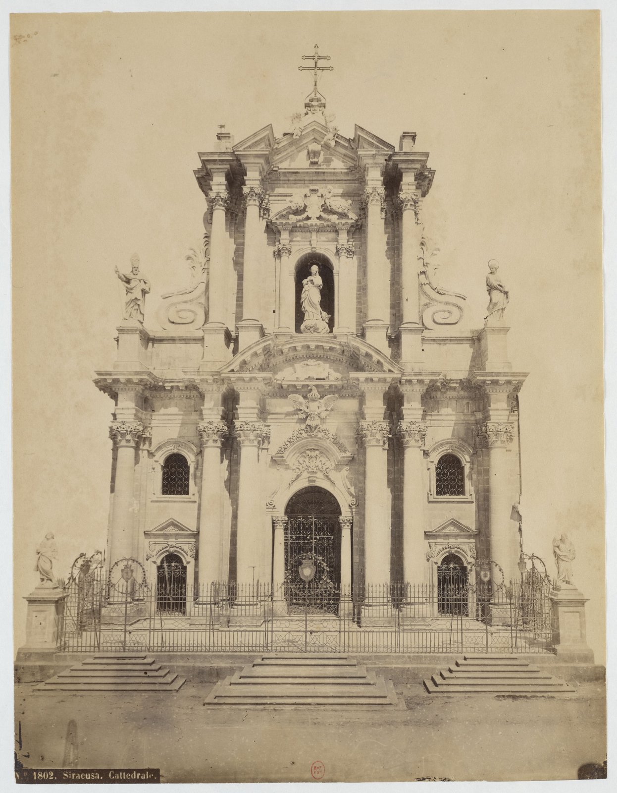 Fotografia d'epoca in tonalità seppia della facciata del Duomo di Siracusa vista frontalmente: due ordini di colonne, portale centrale chiuso da una cancellata in ferro battuto, statua della Vergine nella nicchia del secondo ordine, statue sulle balaustre laterali, croce in ferro sul coronamento e davanti la gradinata vuota con la cancellata del sagrato. In basso a sinistra la didascalia stampata 1802. Siracusa. Cattedrale.