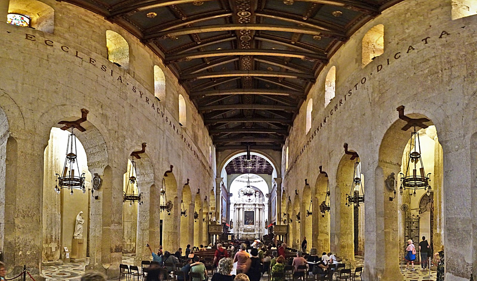 La navata centrale del Duomo di Siracusa vista dall'ingresso verso l'altare maggiore: due file di arcate a tutto sesto aperte nel muro di pietra antica, lampade pensili in ferro battuto, un'iscrizione latina a grandi lettere che corre sopra le arcate, il soffitto ligneo scuro con travi a vista, pannelli dipinti e decorazioni dorate e, in fondo, l'altare barocco bianco illuminato.