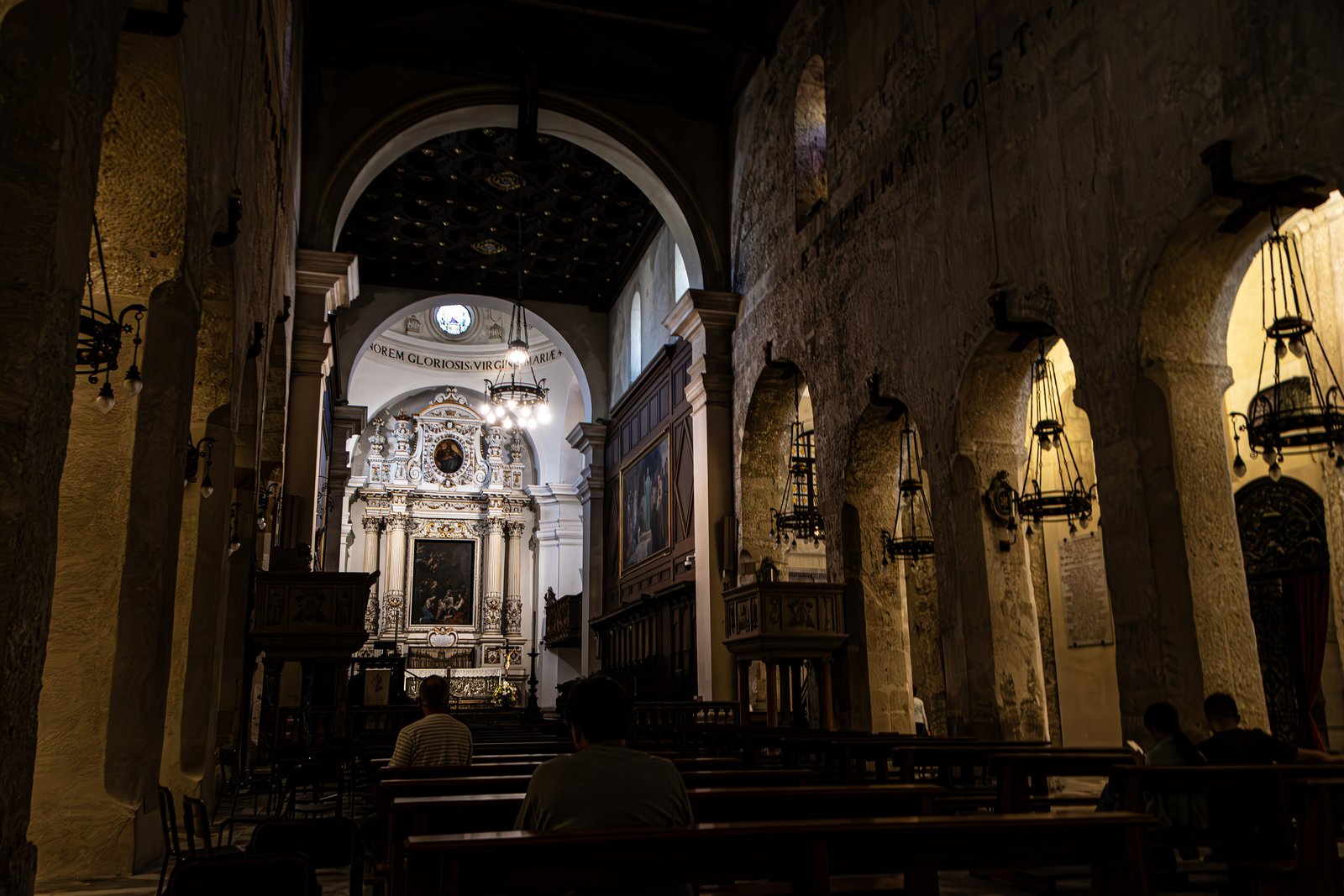 Interno in penombra del Duomo di Siracusa ripreso da una navata laterale: massicci pilastri in pietra grezza ricavati nel muro di cella del tempio greco, arcate profonde con lampade a sospensione, banchi di legno con alcuni fedeli seduti e, in fondo, un altare barocco bianco sotto un arco con iscrizione latina.