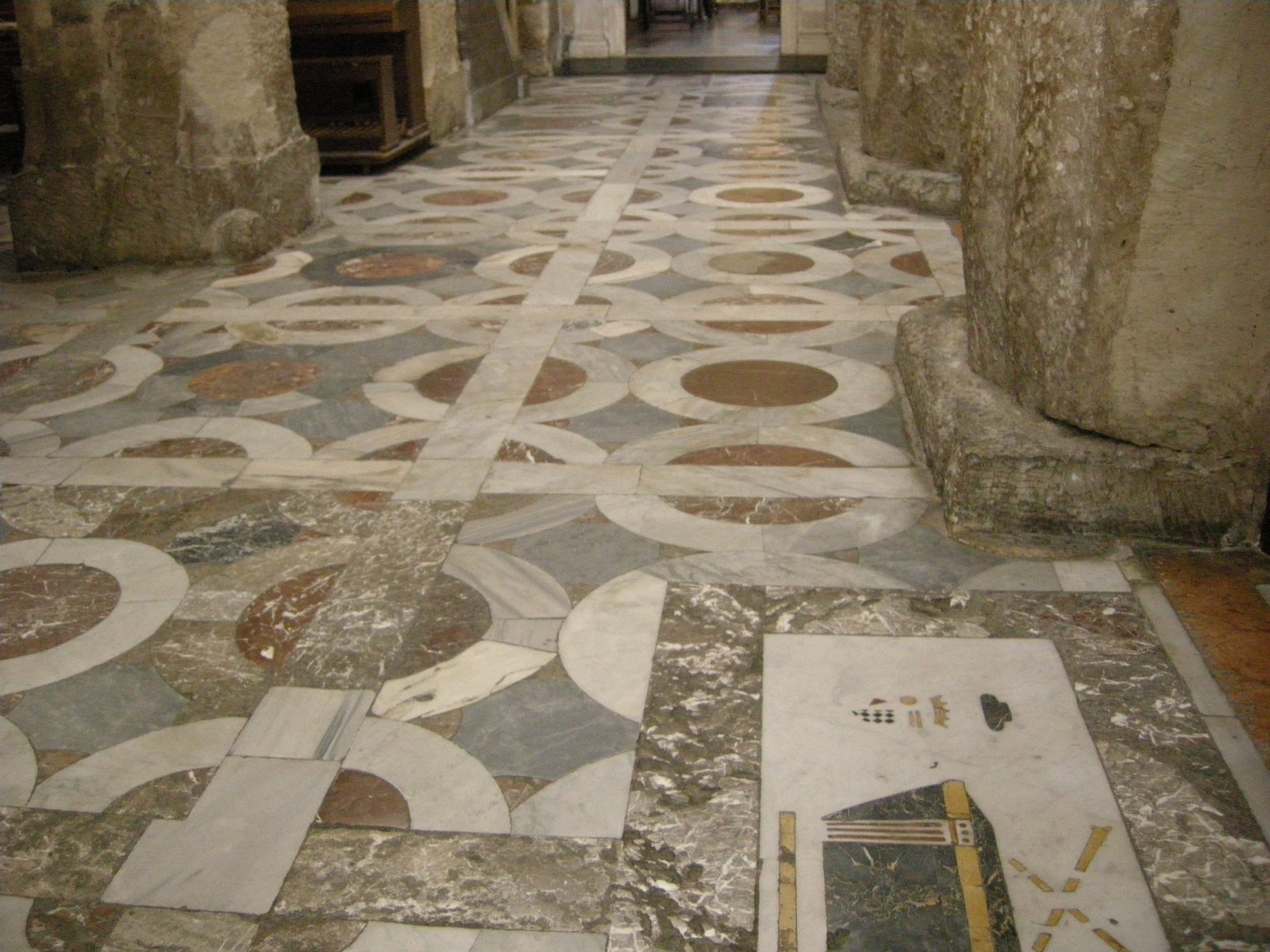 Il pavimento del Duomo di Siracusa ripreso dall'alto in una navata laterale: grandi lastre di marmo bianco, grigio e rosso composte in un disegno geometrico di cerchi e quarti di cerchio, attraversato da una fascia diritta più chiara; a destra la base sagomata di un pilastro in pietra antica e in basso una lastra sepolcrale intarsiata con uno stemma.
