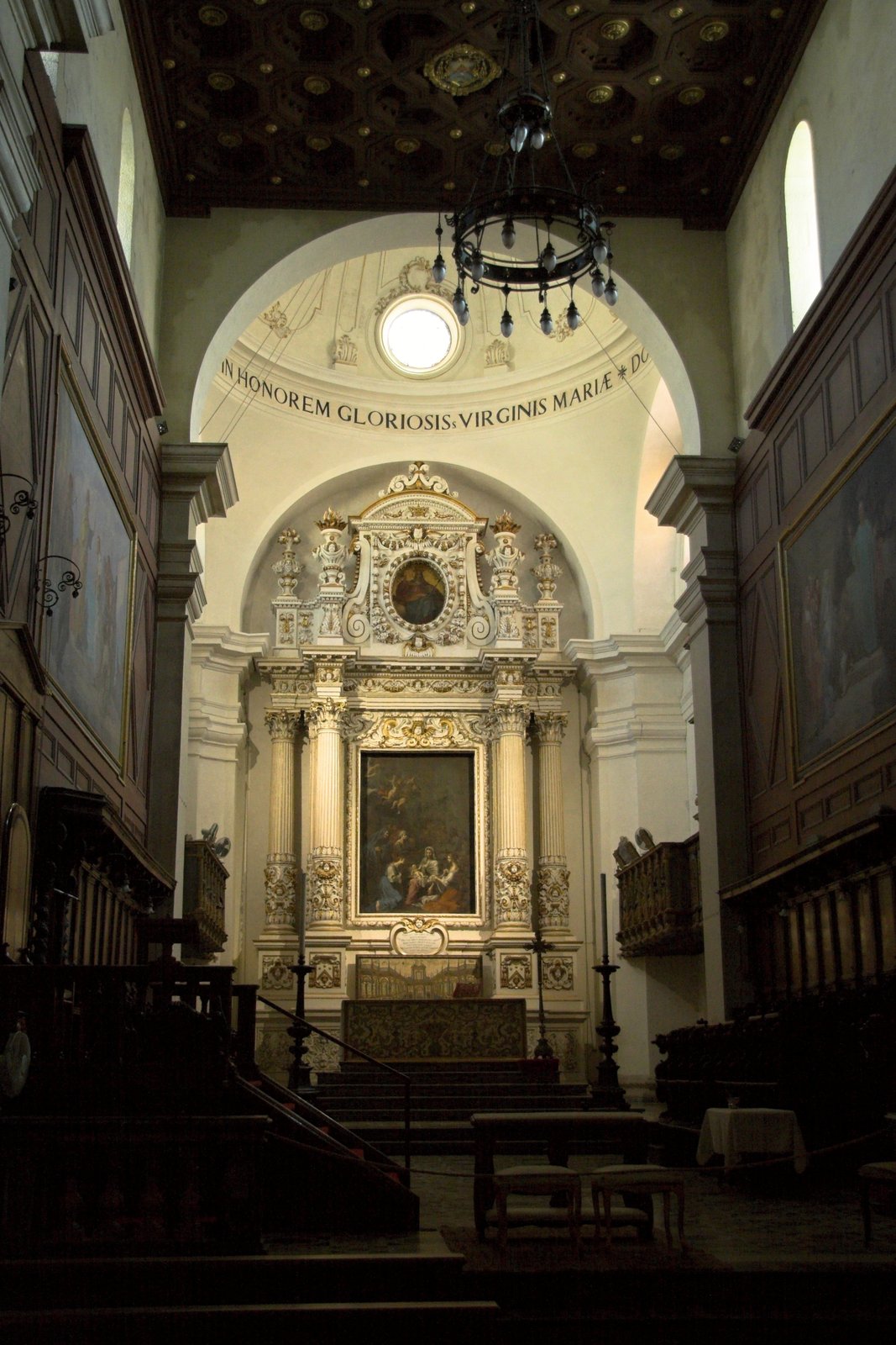 Il presbiterio del Duomo di Siracusa: un grande arco a tutto sesto con iscrizione latina dedicata alla Vergine Maria, un oculo circolare sopra l'arco, un lampadario in ferro battuto appeso al centro, l'altare maggiore barocco in marmo chiaro con colonne e pala dipinta, il coro ligneo ai lati e in alto il soffitto a cassettoni con rosoni dorati.