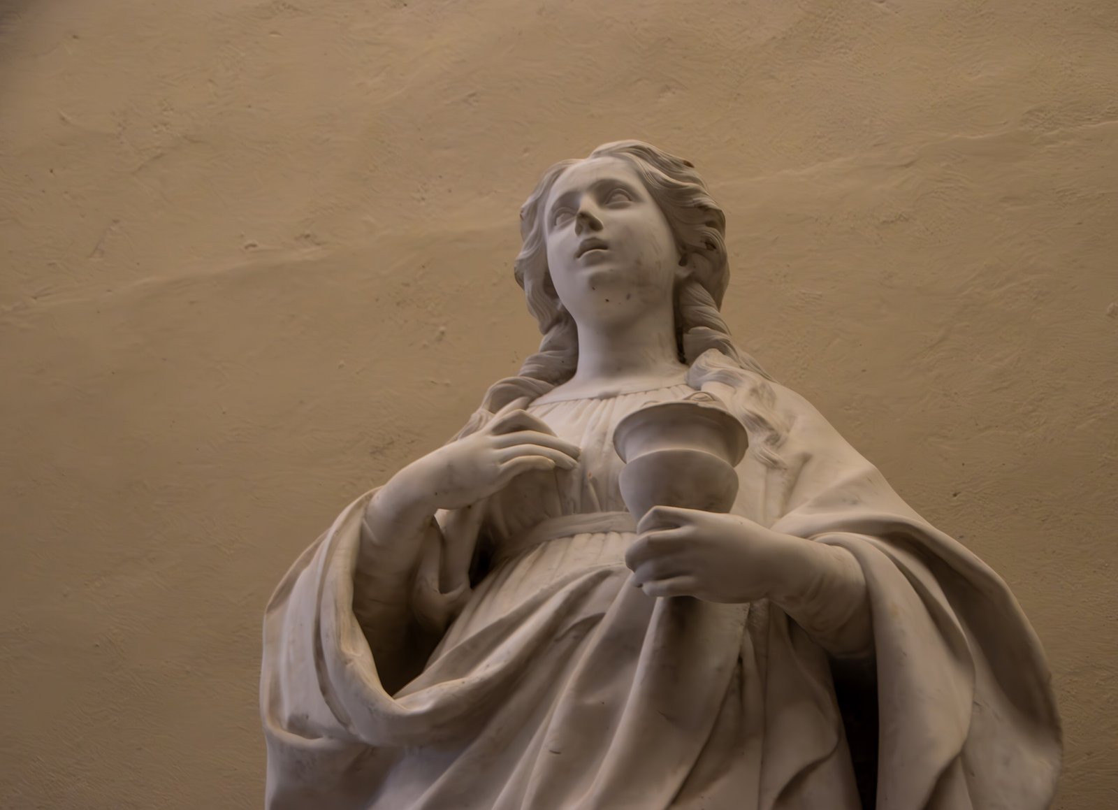Statua marmorea di Santa Lucia ripresa dal basso su una parete color sabbia: lunghi capelli ondulati scendono sulle spalle, il volto è rivolto verso l'alto, un ampio panneggio avvolge il corpo, la mano sinistra regge una coppa e la destra è accostata al petto.